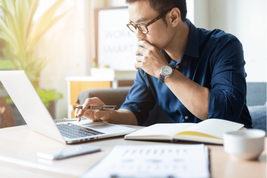 A man with glasses intently working on his laptop, surrounded by office plants and stationery.